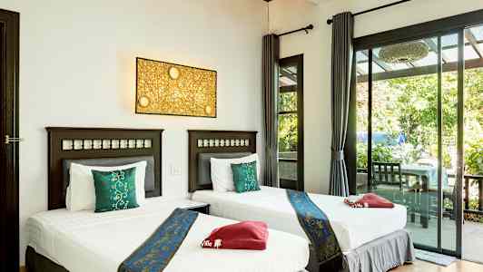 To enkeltsenge med hvidt sengetøj og blå dekorative sengetæpper står side om side i et lyst rum med glasdøre, der åbner ud til en terrasse med udsigt til grønne omgivelser. (AI-genereret tekst)