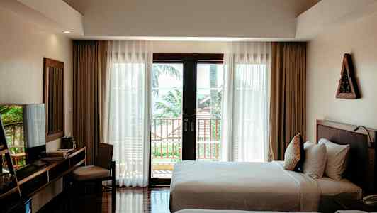 Et hotelværelse med to senge, et skrivebord med stol, og store vinduer med udsigt til en altan og træer udenfor. (AI-genereret tekst)