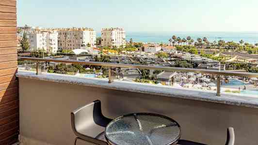 Udsigt fra en altan med bord og stole over en pool og bygninger mod havet. (AI-genereret tekst)