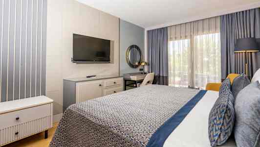 Hotelværelse med moderne indretning, inklusive seng med blå detaljer, skrivebord, TV og store vinduer med gardiner. (AI-genereret tekst)