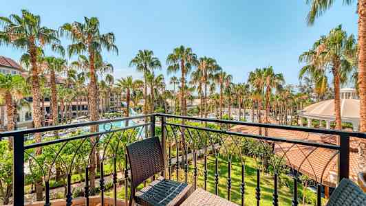 Udsigt fra en altan over en have med palmetræer og bygninger i baggrunden. (AI-genereret tekst)