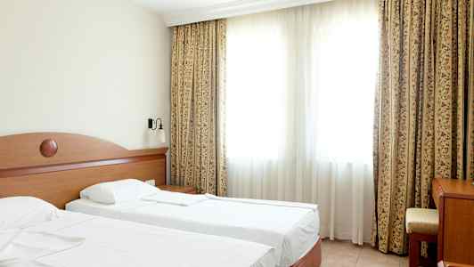 Et hotelværelse med to enkeltsenge, et bord og en stol foran et stort vindue med gardiner. (AI-genereret tekst)