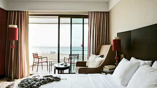 Et hotelværelse med en stor seng, en lænestol og en altan med udsigt over havet. (AI-genereret tekst)