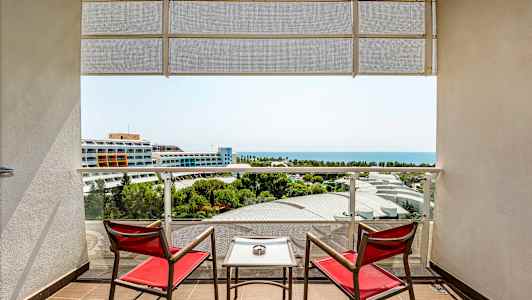 To røde stole og et bord på en altan med udsigt over havet og hotelbygninger. (AI-genereret tekst)