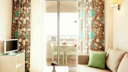 Sofa med grøn pude, TV og et bord med vase foran. Blomstret gardiner og glasdør til en terrasse med bord og stole. (AI-genereret tekst)