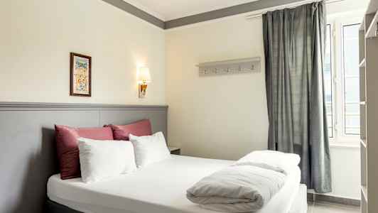 Billede af et hotelværelse med en dobbeltseng, dekoreret med sengetøj i hvidt og rødt samt en lys grå sengegavl. (AI-genereret tekst)