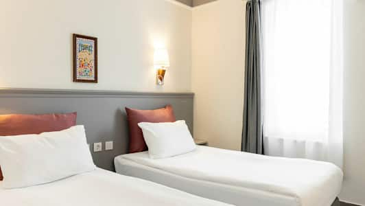 To senge med hvidt sengetøj og røde puder står i et rum med lyse vægge og en væglampe. Et vindue med hvide gardiner er til højre. (AI-genereret tekst)