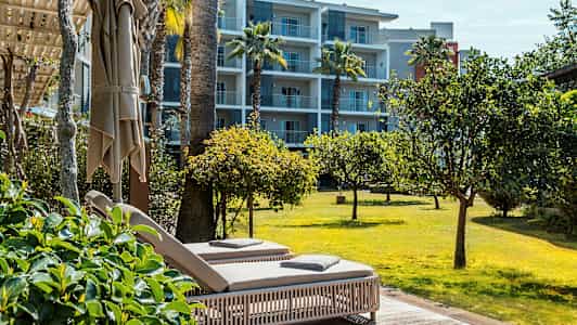 To solsenge på en terrasse med udsigt over en græsplæne og træer, med et lejlighedshotel i baggrunden. (AI-genereret tekst)