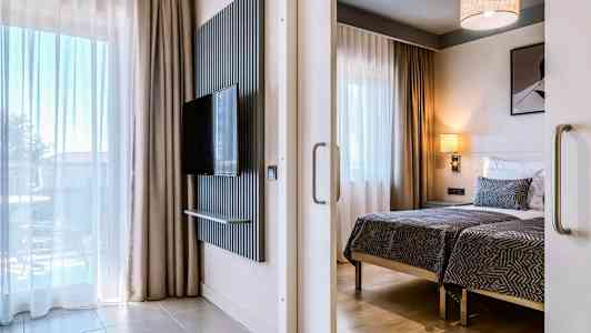 Moderne hotelværelse med to enkeltsenge, et fladskærms-tv på væggen og store vinduer med gardiner. (AI-genereret tekst)