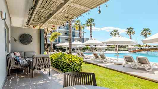 En udendørs veranda med siddepladser og udsigt over en pool med liggestole og parasoller. (AI-genereret tekst)