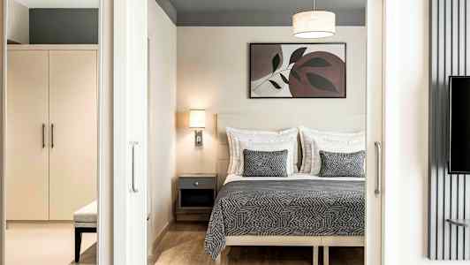 Et soveværelse med en dobbeltseng med gråt sengetæppe og matchende puder, en væglampe og et maleri over sengen. Et skab og bænk ses i forgrunden. (AI-genereret tekst)