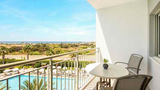 Udsigt fra en altan med stole og bord over et poolområde med liggestole og palmer samt videre mod et åbent landskab og havet. (AI-genereret tekst)