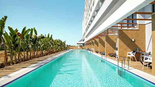 Lang swimmingpool med klart blåt vand ved siden af en bygning med private terrasser og liggestole, omgivet af banantræer. (AI-genereret tekst)