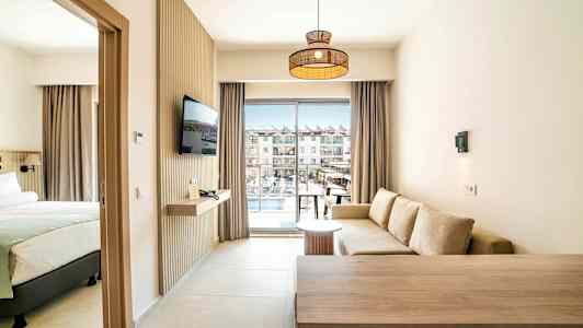 Et hotelværelse med siddeområde, seng og TV samt balkon med udsigt over poolområdet. (AI-genereret tekst)