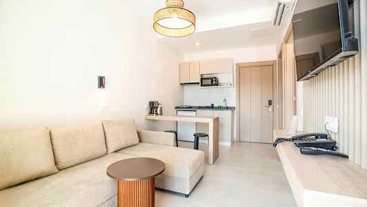 Et hotelværelse med en beige sofa, et lille bord, et køkken og et vægmonteret TV. (AI-genereret tekst)