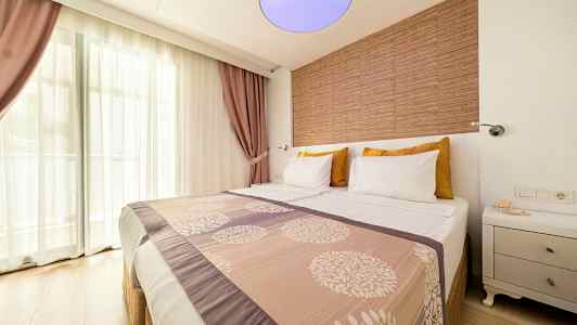 Hotelværelse med en dobbeltseng, dekoreret med mønstret sengetæppe og puder. Vindue med gennemsigtige gardiner og lilla gardiner i baggrunden. (AI-genereret tekst)