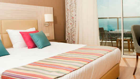 Hotelværelse med en dobbeltseng, farverige puder og havudsigt fra altanen. (AI-genereret tekst)