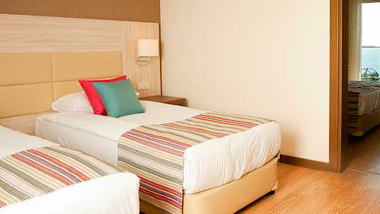 To enkeltsenge med stribede sengetæpper og farverige puder, i et rum med trægulv og lyse vægge. (AI-genereret tekst)