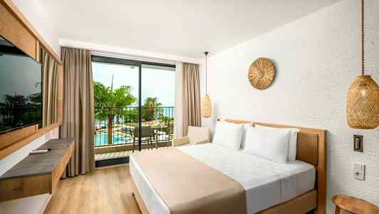 Et hotelværelse med en stor seng og en balkon med udsigt over en pool og palmer. (AI-genereret tekst)