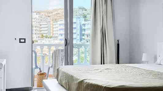 Udsigt fra et hotelværelse med seng og en åben balkondør. Balkon med stole og udsigt over bygninger. (AI-genereret tekst)