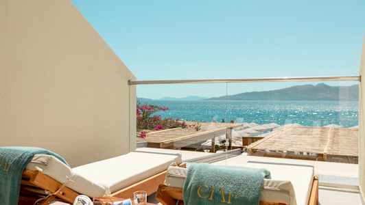 En liggestol med et grønt håndklæde på en balkon med glasrækværk, med udsigt over havet og en strand med parasoller. (AI-genereret tekst)