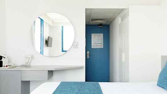 Et hotelværelse med en blå dør, et rundt spejl, et TV og en seng med blå detaljer. (AI-genereret tekst)