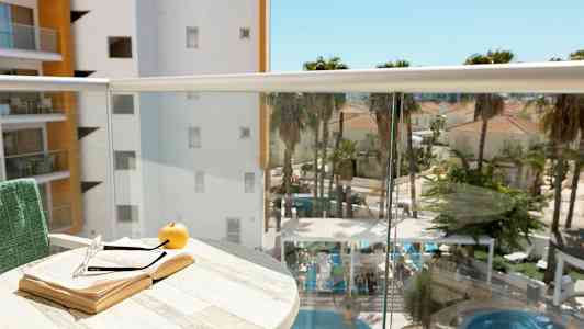 En altan med en åben bog, et par briller og et æble på et bord. I baggrunden ses en pool med omkringliggende palmer og bygninger. (AI-genereret tekst)