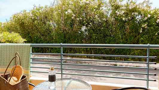 Udsigt fra en altan med et glasbord, stole og frodig beplantning i baggrunden. (AI-genereret tekst)