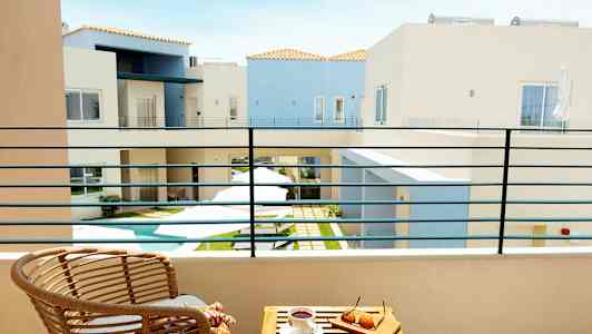 Udsigt fra en balkon til en gårdhave med en lille pool omgivet af lyse bygninger. På balkonen er der en stol og et bord med solbriller og en kaffekop. (AI-genereret tekst)