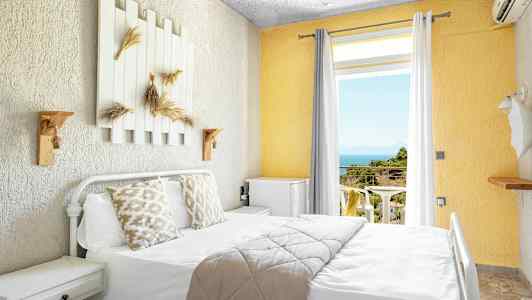 Lyst hotelværelse med seng, dekorerede puder og udsigt til altan med hav og grønt. (AI-genereret tekst)