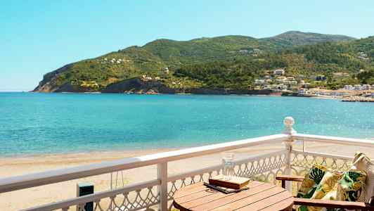 Udsigt fra en balkon mod havet og frodige bakker, med en træstol og et bord. (AI-genereret tekst)