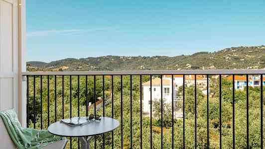 Udsigt fra en balkon med bord og stol over et frodigt landskab med huse og bjerge. (AI-genereret tekst)