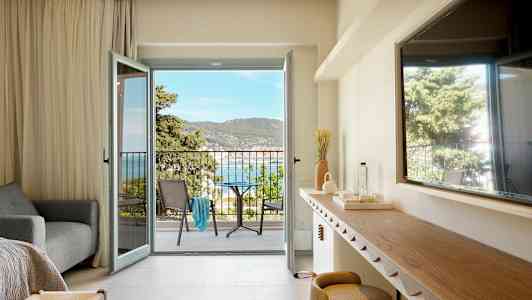 Et hotelværelse med åben altandør, der viser udsigt over havet og omkringliggende beplantning. (AI-genereret tekst)