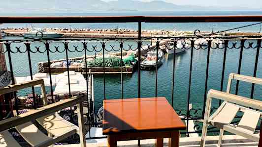 Udsigt fra en altan med bord og stole mod en havn med både og en stenmole. (AI-genereret tekst)