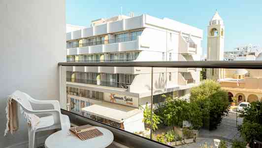 Udsigt fra en altan med et bord og en stol, med en bygning og et kirketårn i baggrunden. (AI-genereret tekst)