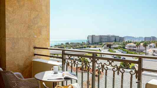 En balkon med et rundt bord, to stole og udsigt over bygninger, veje og havet. (AI-genereret tekst)