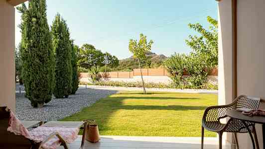 Udsigt fra en terrasse med stole og bord, græsplæne og træer i baggrunden. (AI-genereret tekst)