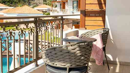 En altan med to siddepladser, et lille bord og en flot udsigt over en swimmingpool og bygninger. (AI-genereret tekst)