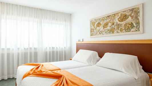 Et hotelværelse med to senge beklædt med hvide sengelagner og orange dekorative sjaler, en vægdekoration over sengene og gardiner foran et stort vindue. (AI-genereret tekst)