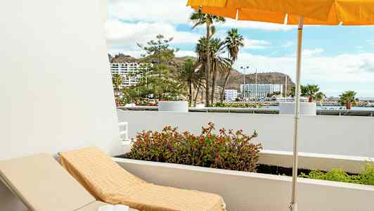 Liggestol med håndklæde under parasol på en altan med udsigt til palmer og bygninger. (AI-genereret tekst)