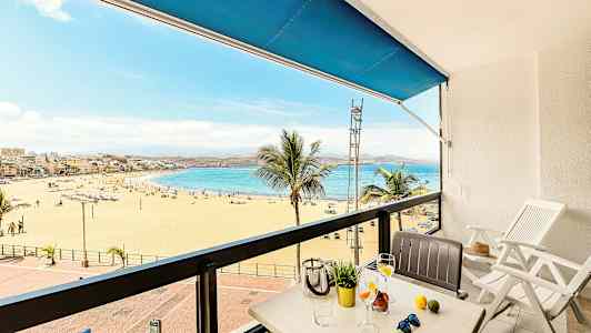 Udsigt fra en altan mod en sandstrand og hav, med bord og stole i forgrunden. (AI-genereret tekst)