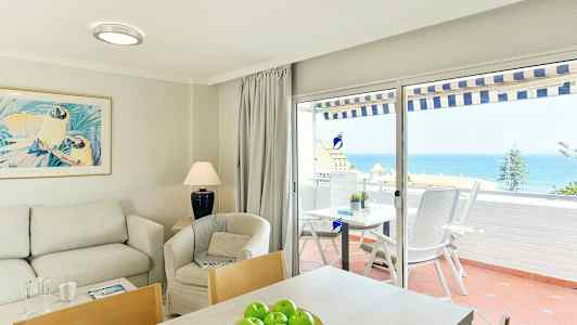 Interiør af en stue med sofa, lænestol og bord, samt udsigt over havet gennem glasdøre til en terrasse. (AI-genereret tekst)