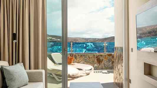 En udsigt fra et rum mod en terrasse med liggestole og glasrækværk, landskab og himmel i baggrunden. (AI-genereret tekst)