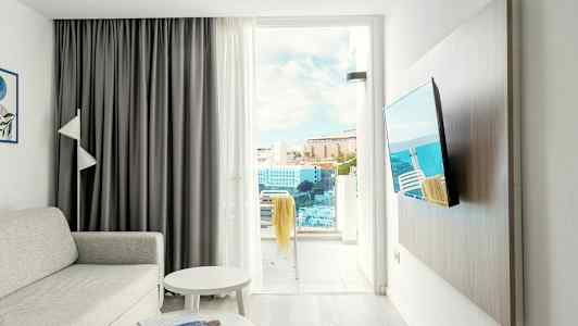 En stue med sofa, bord, TV og en åben altandør med udsigt over bygninger og havet. (AI-genereret tekst)