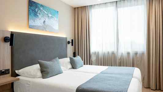 Et hotelværelse med to senge dækket med blå sengetøj og to puder, foran vinduer med lyse gardiner. (AI-genereret tekst)