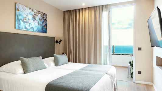 Et hotelværelse med en dobbeltseng, beige gardiner og havudsigt gennem en glasdør. (AI-genereret tekst)