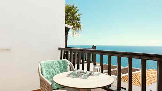 Udsigt fra en altan over havet med en stol, et bord og et grønt tæppe. (AI-genereret tekst)