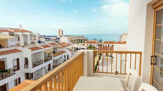 Udsigt fra en balkon med et bord og stole over hvide bygninger med røde tage og havet i baggrunden. (AI-genereret tekst)