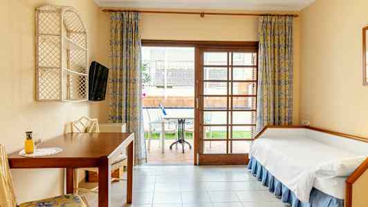 Et hotelværelse med en enkeltseng, bord og stole samt udsigt mod en terrasse med møbler. (AI-genereret tekst)