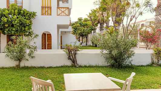 Havemøbler med bord og stole foran en grøn græsplæne og hvide bygninger. (AI-genereret tekst)